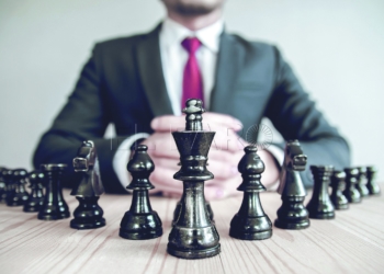 Retro style image of a businessman with clasped hands planning strategy with chess figures on an old wooden table.