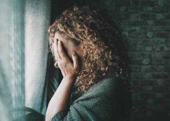 Sad and worried woman side portrait at home in the dark looking outside the window. Worried and lonely female alone indoor. Concept of end of relationship and unsuccessful emotion. Lady touching face