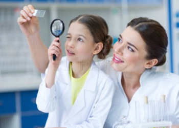 Woman teacher and girl student scientists looking at glass microscope slide through magnifier