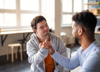 A young happy man with Down syndrome with his mentoring friend celebrating success indoors at school.