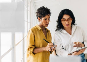 Two business women having a discussion, they're standing in an office and using a tablet. Professional women making a to do list as part of their project planning.